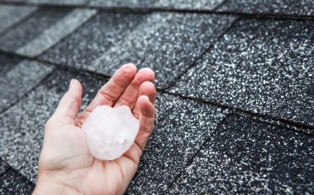 A large piece of hail in a person's hand over  some roofing shingles.